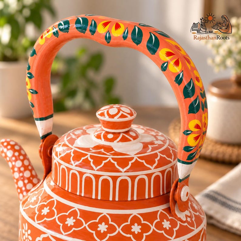 May include: An orange metal teapot featuring white floral patterns on the body and lid, with a handle decorated in green leaves and yellow flowers. The text RajasthanRoots is visible in the upper right corner with a small graphic illustration.
