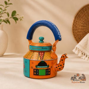 Puede incluir: Una tetera de metal pintada a mano con una base naranja vibrante y un motivo arquitectónico en verde y negro. El asa es azul con detalles florales blancos y la tapa es de color verde azulado. El texto Rajasthan Roots está impreso.