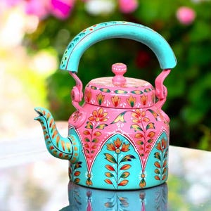 Può includere: Una teiera dipinta a mano rosa e turchese con disegni floreali. La teiera presenta un manico e un beccuccio turchese, con dettagli rosa e oro. Il design include fiori arancioni e rossi e foglie verdi.