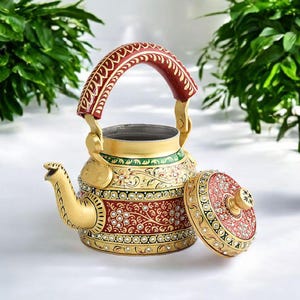 May include: Decorative gold-colored teapot with a red handle and accents. The teapot features intricate floral patterns in red and green, embellished with small, sparkling stones. The lid is off, revealing the interior.