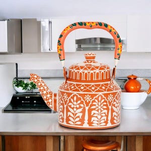 May include: A decorative orange and white patterned tea kettle with a matching lid and handle. The handle is decorated with yellow, green, and orange floral designs. The kettle is sitting on a stainless steel countertop.