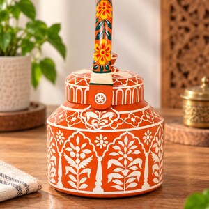 May include: A bright orange metal teapot featuring intricate white floral and leaf patterns across its body. The handle is decorated with yellow and orange flowers on a green background. The lid has a matching orange and white design.