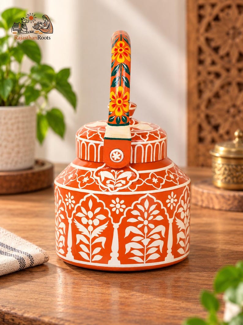 May include: A bright orange metal teapot featuring intricate white floral and leaf patterns across its body. The handle is decorated with yellow and orange flowers on a green background. The lid has a matching orange and white design.