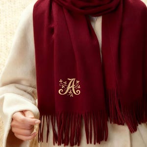 Peut inclure: Écharpe bordeaux avec un « A » brodé en or et des franges. L'écharpe est enroulée autour du cou et des épaules. La personne porte un manteau de couleur crème.