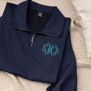 May include: A navy blue quarter-zip sweatshirt with a teal monogram. The long-sleeved top features a collared neckline and ribbed cuffs. The monogram is embroidered on the front. The garment is made of a soft material.