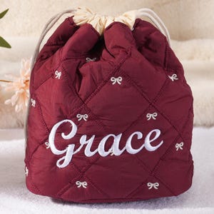 May include: A burgundy quilted drawstring bag with white bow accents and the name "Grace" embroidered in white. The bag has a cream-colored drawstring closure and is likely used for cosmetics or small items.