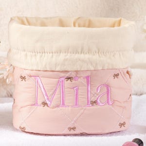 以下が含まれることがあります： ピンク色のキルティング生地の収納バスケット。名前の「Mila」がピンク色で刺繍されています。バスケットはクリーム色の折り返しトップで、小さなリボンアクセントで装飾されています。バスケットは小物を収納するように設計されています。