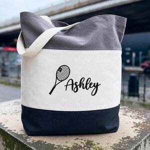 May include: A colour-blocked canvas tote bag featuring a dark grey top section, a white middle panel, and a dark navy blue bottom section. The white panel displays a black graphic of a tennis racket with a ball and the name Ashley in script.