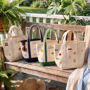May include: Four canvas tote bags with embroidered patches and personalized names. The bags feature various colored bases including natural, navy, and green. Each bag displays unique small icons like cherries, a smiley face, and a ladybug.