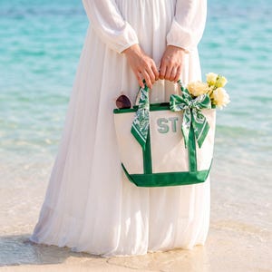 May include: A cream-coloured canvas tote bag with green trim and base, featuring the letters ST in block font. The bag is adorned with a green and white patterned silk scarf tied in a bow on one handle and contains a bouquet of pale yellow roses.