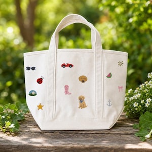 May include: A cream-coloured canvas tote bag featuring various embroidered icons including a red car, a golden retriever, a ladybug, sunglasses, a pink cowboy boot, a watermelon slice, a star, an anchor, a bow, and a small green plant sprig.