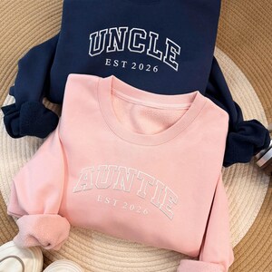 Puede incluir: Dos sudaderas, una azul marino y otra rosa. La sudadera azul marino tiene la palabra "UNCLE" y "EST 2026" impresas en blanco. La sudadera rosa tiene la palabra "AUNTIE" y "EST 2026" impresas en blanco. Ambas sudaderas tienen cuello redondo.
