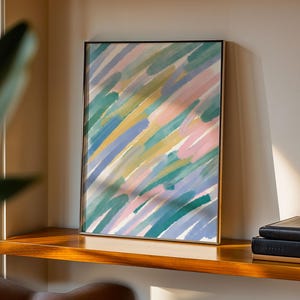 Puede incluir: Una impresión artística enmarcada con pinceladas diagonales en tonos azules, verdes, rosas y amarillos. La obra de arte se exhibe en una estantería de madera, con libros a la derecha.