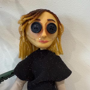 May include: A handmade doll with button eyes, yellow hair, and a black dress. The doll's face is painted with a stitched smile and a small beauty mark. The doll's hair is styled in two braids.