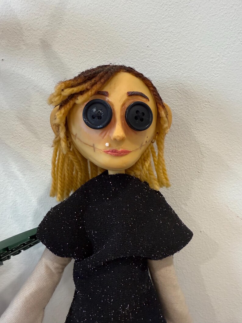 May include: A handmade doll with button eyes, yellow hair, and a black dress. The doll's face is painted with a stitched smile and a small beauty mark. The doll's hair is styled in two braids.