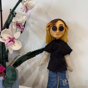 May include: A handmade doll with button eyes, yellow yarn hair, and a stitched face. The doll wears a black glittery top and distressed denim jeans. A Lego orchid plant is in the background.