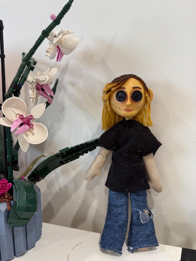 May include: A handmade doll with button eyes, yellow yarn hair, and a stitched face. The doll wears a black glittery top and distressed denim jeans. A Lego orchid plant is in the background.