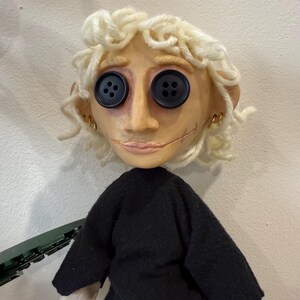 May include: A handmade doll with a pale face, button eyes, and stitched mouth. The doll has curly blonde yarn hair, small gold hoop earrings, and wears a black top. The doll's arms are a light beige color.