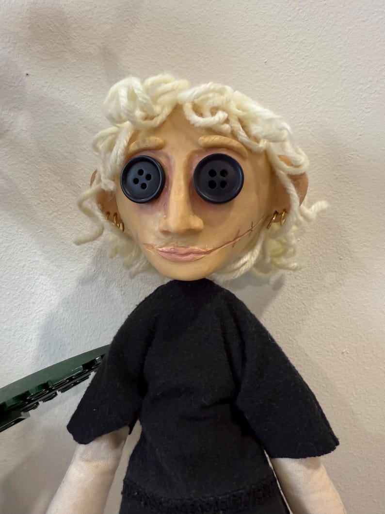 May include: A handmade doll with a pale face, button eyes, and stitched mouth. The doll has curly blonde yarn hair, small gold hoop earrings, and wears a black top. The doll's arms are a light beige color.