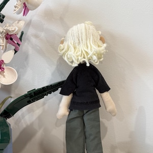 May include: A handmade doll with a black top and olive green pants. The doll has a cream-colored yarn wig and beige fabric hands and feet. The doll is standing in front of a white wall.