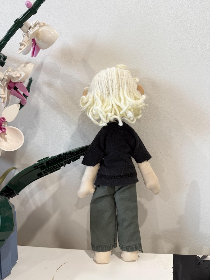 May include: A handmade doll with a black top and olive green pants. The doll has a cream-colored yarn wig and beige fabric hands and feet. The doll is standing in front of a white wall.