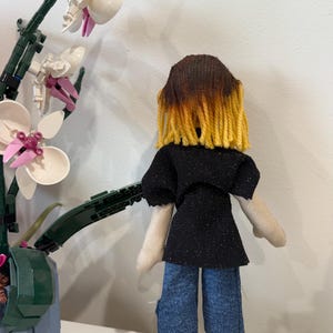May include: A handmade doll with brown and yellow yarn hair, wearing a black glitter top and blue jeans. The doll is positioned in front of a white and pink orchid plant.