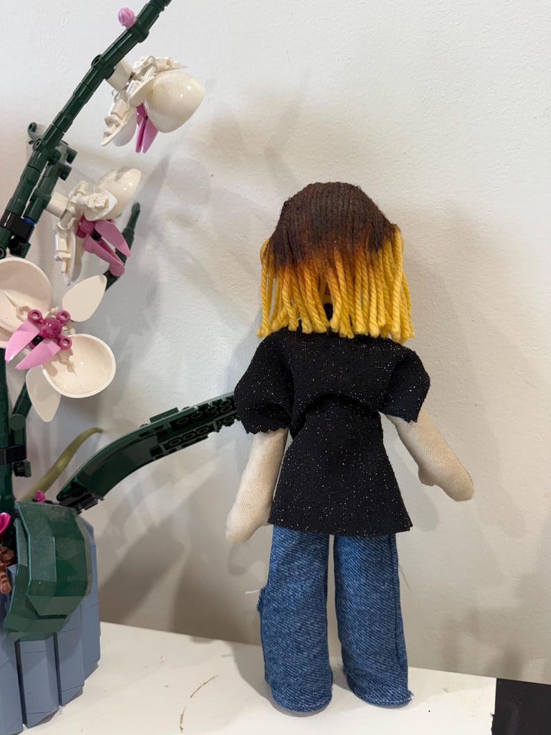 May include: A handmade doll with brown and yellow yarn hair, wearing a black glitter top and blue jeans. The doll is positioned in front of a white and pink orchid plant.