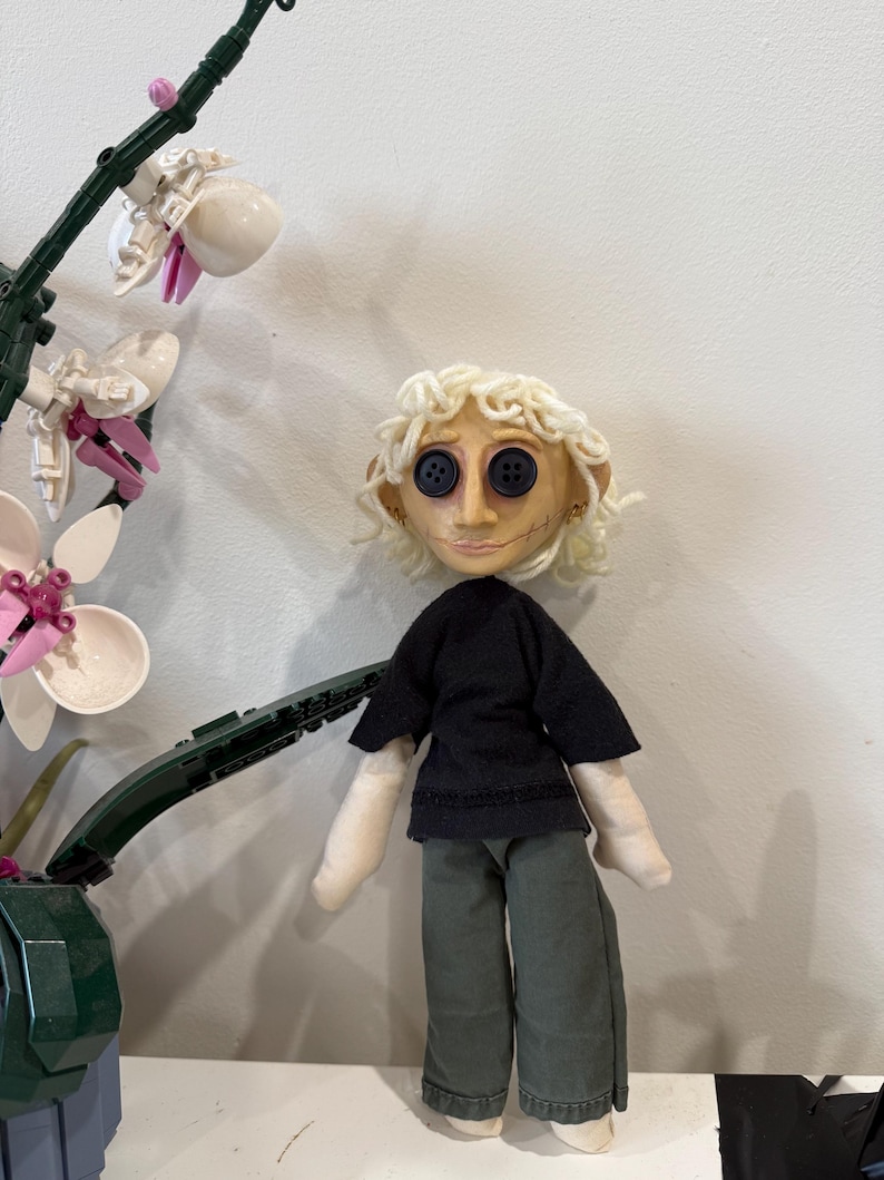 May include: A handmade doll with button eyes, blonde curly hair, and stitched facial features. The doll wears a black top and green pants. A decorative orchid plant is in the background.