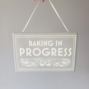 May include: A rectangular light gray hanging sign with white text that reads BAKING IN PROGRESS. The sign features a white border and a decorative white scrollwork pattern at the bottom. It is suspended by a thin white string attached to the top.