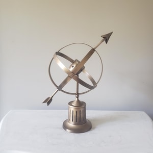 May include: A bronze-toned metal armillary sphere sculpture featuring intersecting rings and a central arrow pointing diagonally. The object is mounted on a cylindrical pedestal base with vertical cutouts, showcasing a metallic finish throughout.