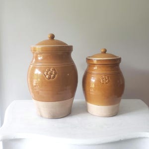 May include: Two matching ceramic jars with lids. The jars are a warm brown color with a cream-colored base. Each jar has a decorative floral design and a rounded lid with a small knob. The jars are likely for kitchen storage.