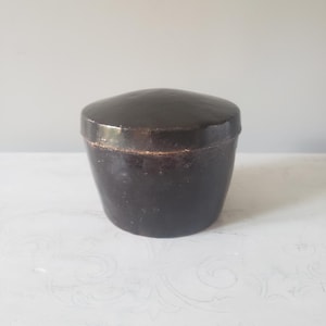 May include: A dark brown, round, lidded container with a smooth, glossy finish. The container features a slightly tapered cylindrical body and a domed lid that fits securely over the top edge, showing subtle signs of wear along the rim.