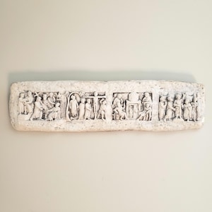 May include: A rectangular, off-white relief sculpture depicting multiple scenes with figures. The scenes appear to be religious or historical, with detailed carvings of people and objects. The sculpture is mounted on a neutral-colored wall.