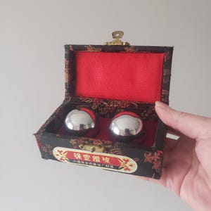 May include: A pair of shiny, silver metal Baoding balls inside a black box with red lining. The box has a gold clasp and Chinese text on the front. The box is decorated with a floral pattern.