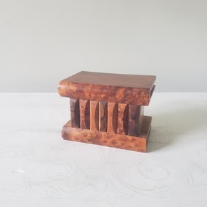 May include: A rectangular wooden box featuring a polished burl wood grain pattern. The box consists of a flat top and base connected by a series of vertical, curved slats that create an open, ribbed appearance along the front and sides of the object.