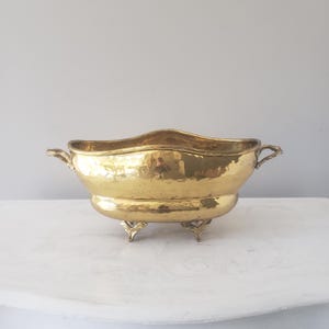 May include: A polished brass bowl with a hammered texture, featuring an elongated oval shape, two decorative side handles, and four ornate feet. The metallic surface has a reflective gold finish with subtle indentations throughout.