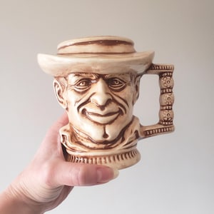 May include: A vintage ceramic mug shaped like a man's head, wearing a hat. The mug is beige with brown accents, featuring a detailed face and a decorative handle. The mug is held by a hand.