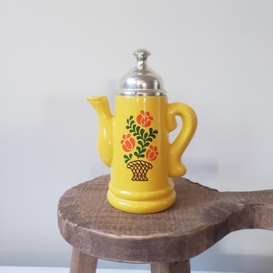May include: A yellow ceramic teapot with a silver lid and a handle. The teapot is decorated with a floral design in orange and green. It is sitting on a small, round, wooden stool.