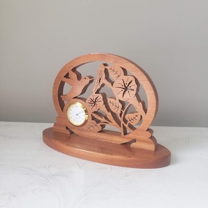 May include: A wooden table clock featuring an oval frame with intricate cutouts of a hummingbird and morning glory flowers. The clock face is circular with a gold-toned rim, white background, black numerals, and black hands.