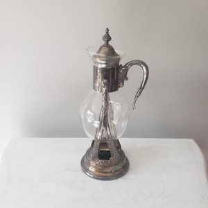 May include: A vintage silver-plated coffee maker with a clear glass carafe. The coffee maker has an ornate design with a handle and a decorative lid. The base and the top are silver-plated, and the carafe is a clear glass.