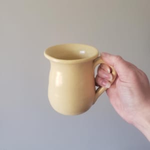 May include: A pale yellow ceramic mug with a rounded body and a handle. The mug has a slightly flared rim and is held by a hand against a neutral background. The mug is a simple, classic design.