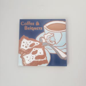 May include: A white ceramic mug with a black handle and the text "But First, Coffee" in a bold, black font. The mug is filled with a dark liquid, likely coffee, and sits on a light-colored surface.