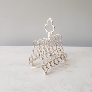 May include: A silver-toned metal toast rack featuring a series of six arched, scalloped wire dividers. The structure includes a central vertical handle with a decorative clover-shaped top and rests on four small, rounded ball-shaped feet.