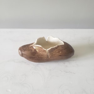 May include: A handcrafted ceramic bowl with a unique, organic shape. The exterior is a mottled brown, while the interior is a creamy white. The bowl has a split design and is suitable for decorative use or holding small items.