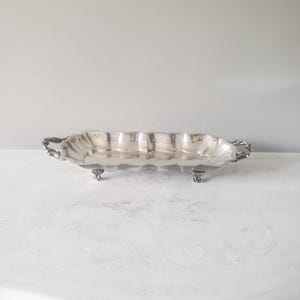 May include: A rectangular silver-toned serving tray with a scalloped edge and a polished metallic finish. The tray features two ornate handles on the sides and is supported by four small, decorative feet, providing an elevated presentation.