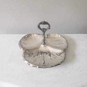 May include: A silver-colored, three-sectioned serving dish with a decorative handle. The dish has a scalloped edge and a leaf-like shape. The handle is ornate and adds to the vintage aesthetic. The dish is likely made of metal.