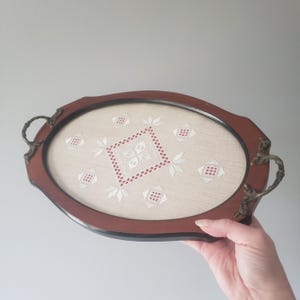 May include: An oval serving tray with a dark brown wooden frame and ornate brass handles. The center features a beige fabric inlay decorated with white and red geometric embroidery, including a central diamond pattern and smaller motifs.