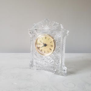 May include: A clear, cut-glass mantel clock featuring a gold-toned circular face with black Roman numerals and hands. The clock is encased in a decorative, faceted crystal frame with intricate geometric patterns and a pointed top design.