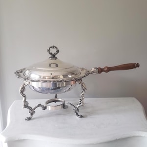 May include: A silver chafing dish with a lid and ornate detailing. The dish has a wooden handle and sits on decorative legs. The lid features a heart-shaped handle. The chafing dish is designed for serving food.