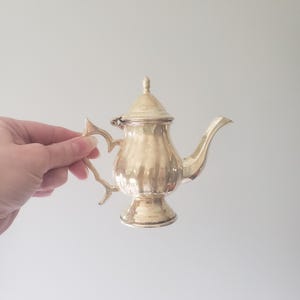 May include: A small, ornate silver teapot with a lid and a curved spout. The teapot has a decorative handle and a ribbed body. The teapot is held by a hand against a plain background. The teapot is likely vintage or antique.